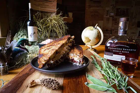 Gourmet sandwich with melted cheese and ingredients on a wooden platter, surrounded by a bottle of Woodford Reserve bourbon, a glass, a wine bottle, and decorative herbs, reflecting the casual dining atmosphere of The Duck Blind Grille.
