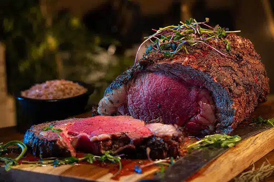 Succulent prime rib roast garnished with herbs, served on a wooden cutting board alongside a bowl of seasoning, highlighting the inviting dining experience at The Duck Blind Grille.