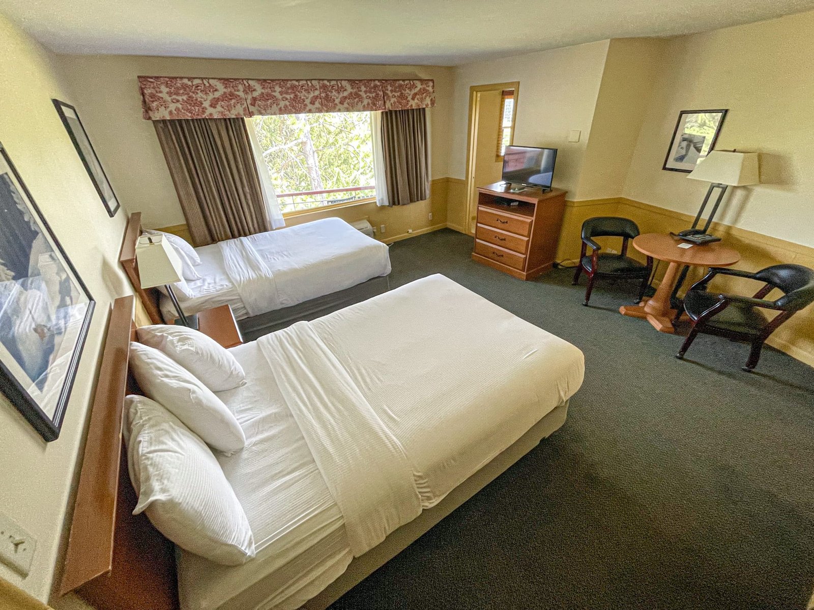 Hotel room at Blue Spruce Lodge featuring two beds, a sitting area with a table and chairs, flat screen TV, and window with natural light.
