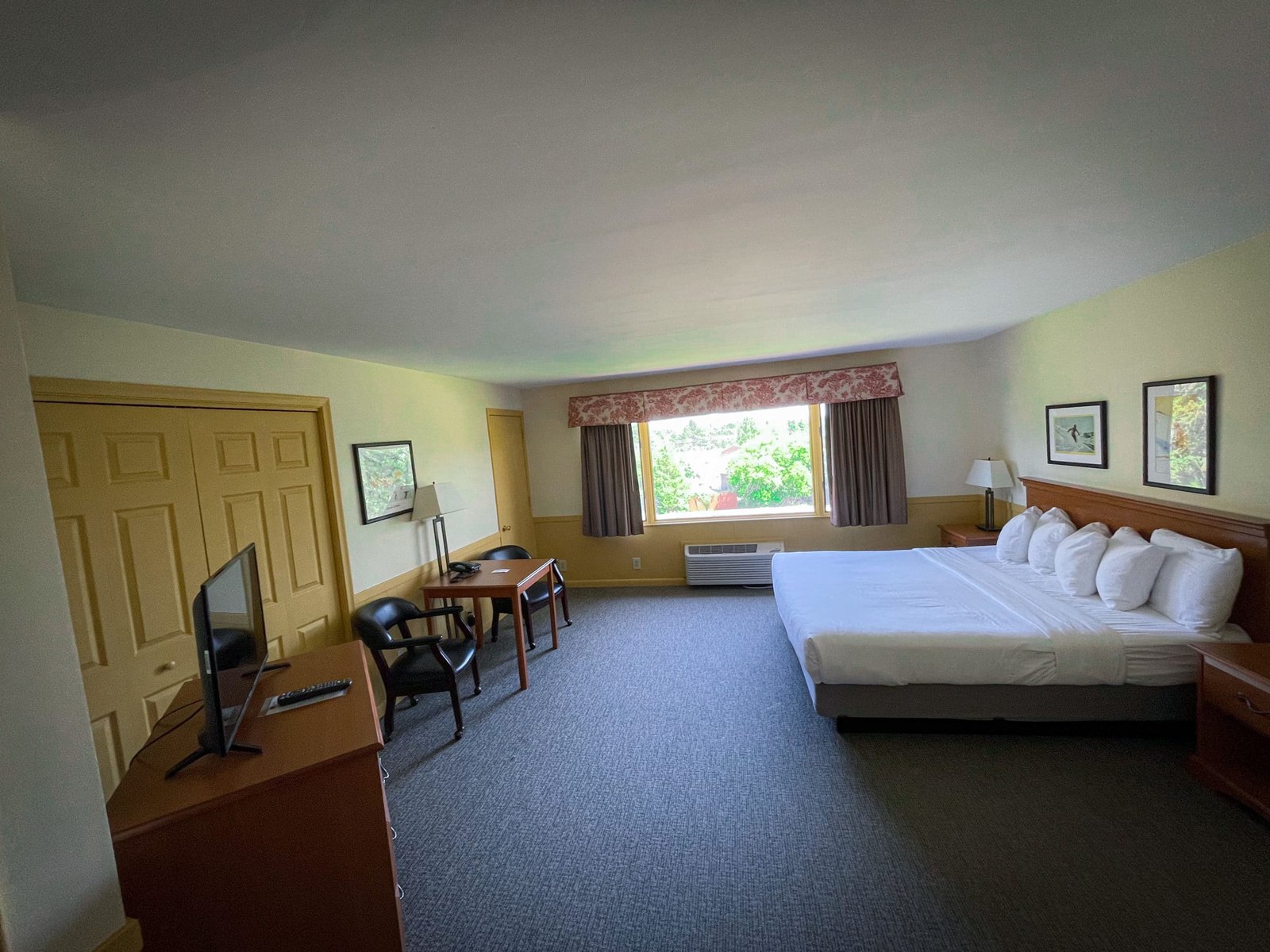 Spacious Blue Spruce Lodge hotel room featuring a king-size bed with plush pillows, sitting area with table and chairs, flat-screen TV, and large windows offering natural light.