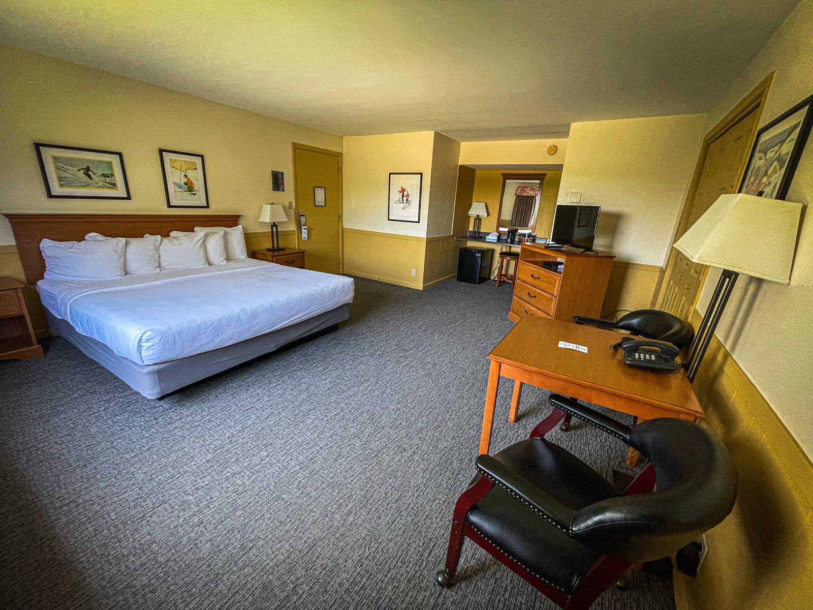 Hotel room at Blue Spruce Lodge featuring a king-size bed, desk with chairs, flat-screen TV, and warm lighting for a comfortable stay.