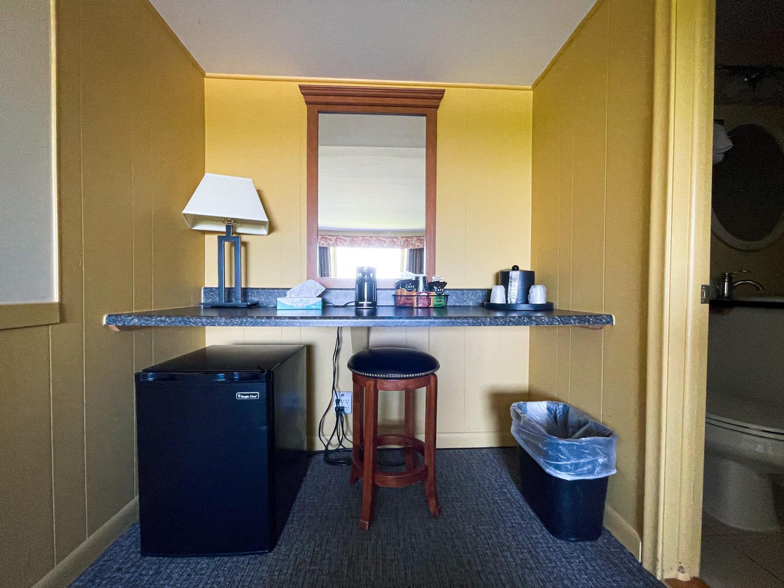 Hotel room corner with a bar-style seating area, black mini fridge, lamp, coffee supplies, and a wastebasket, reflecting the comfort and amenities of Blue Spruce Lodge.