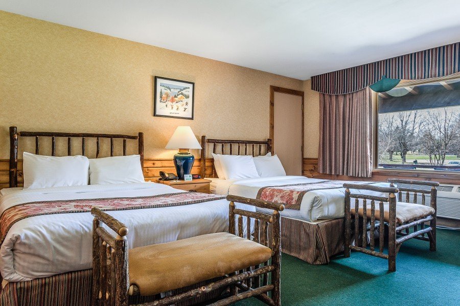 Hotel room at Blue Spruce Lodge featuring two beds, rustic decor, plush bedding, and a cozy sitting area with a lamp and artwork, showcasing comfortable accommodations for travelers and families.