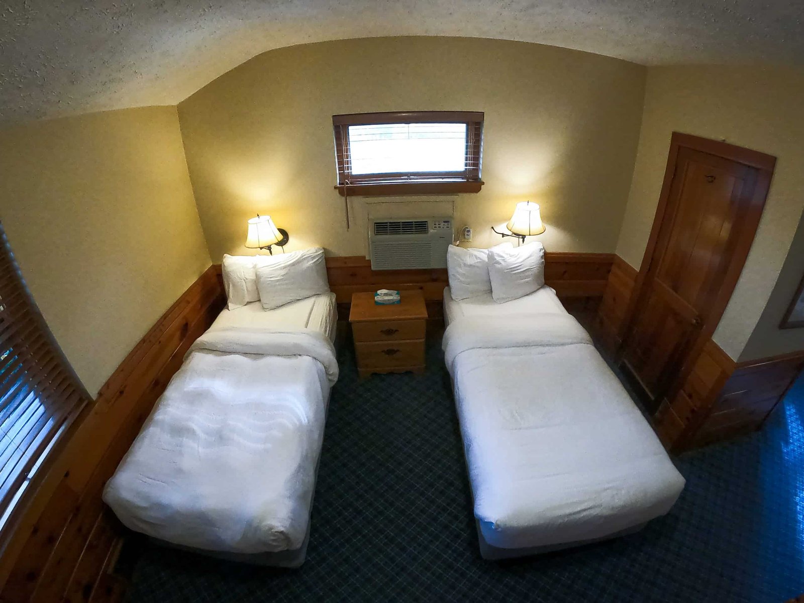 Spacious chalet bedroom with two twin beds, bedside lamp, wooden furniture, and air conditioning, showcasing cozy accommodations at Otsego Resort.