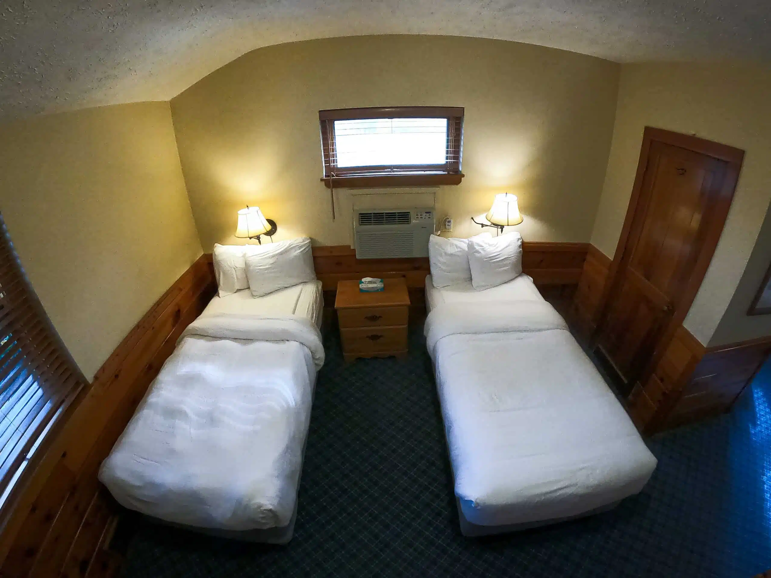 Spacious chalet bedroom with two twin beds, bedside lamp, wooden furniture, and air conditioning, showcasing cozy accommodations at Otsego Resort.