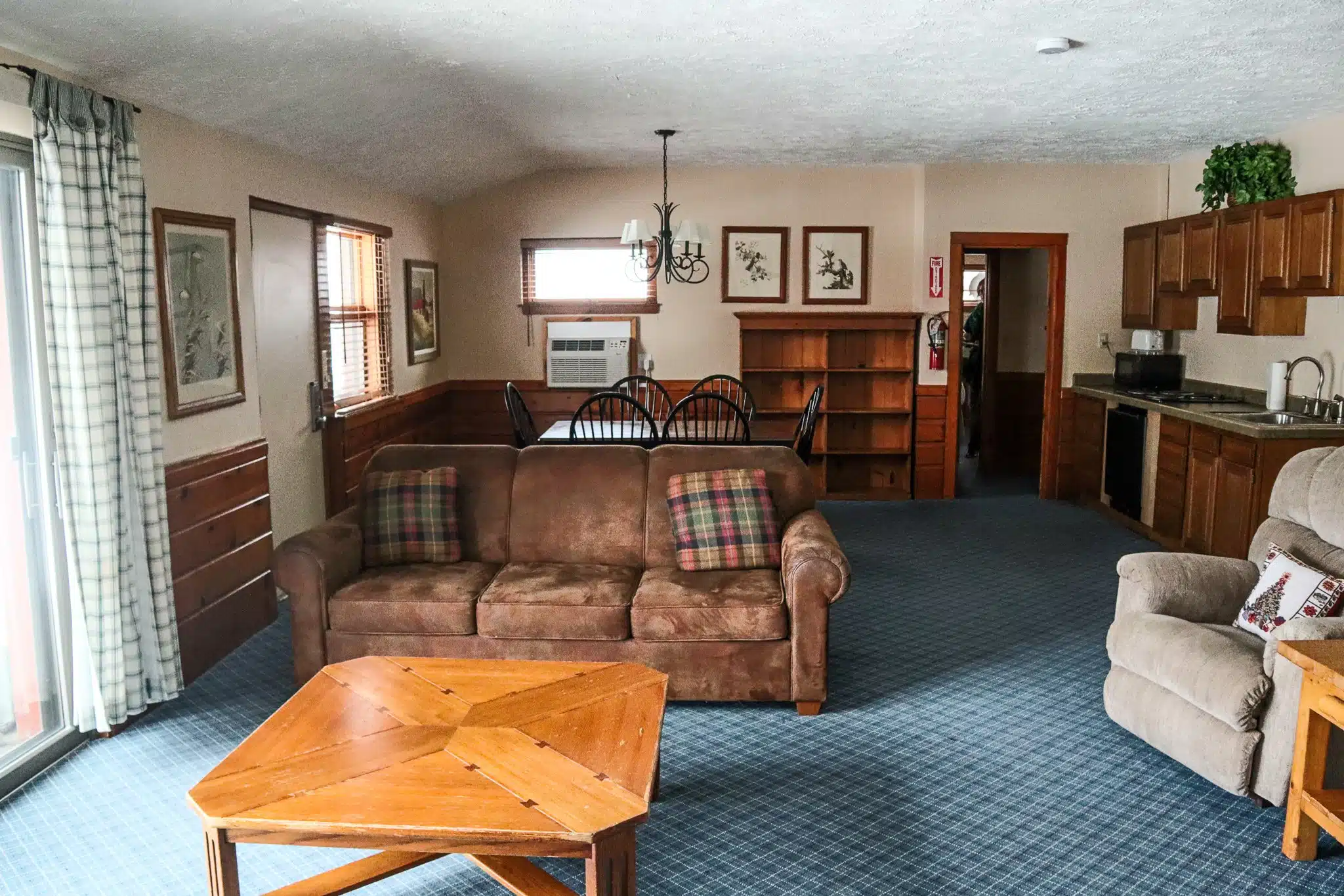 Spacious chalet living room featuring a brown sofa with plaid cushions, wooden coffee table, dining area, kitchenette, and large windows, designed for comfort at Otsego Resort.