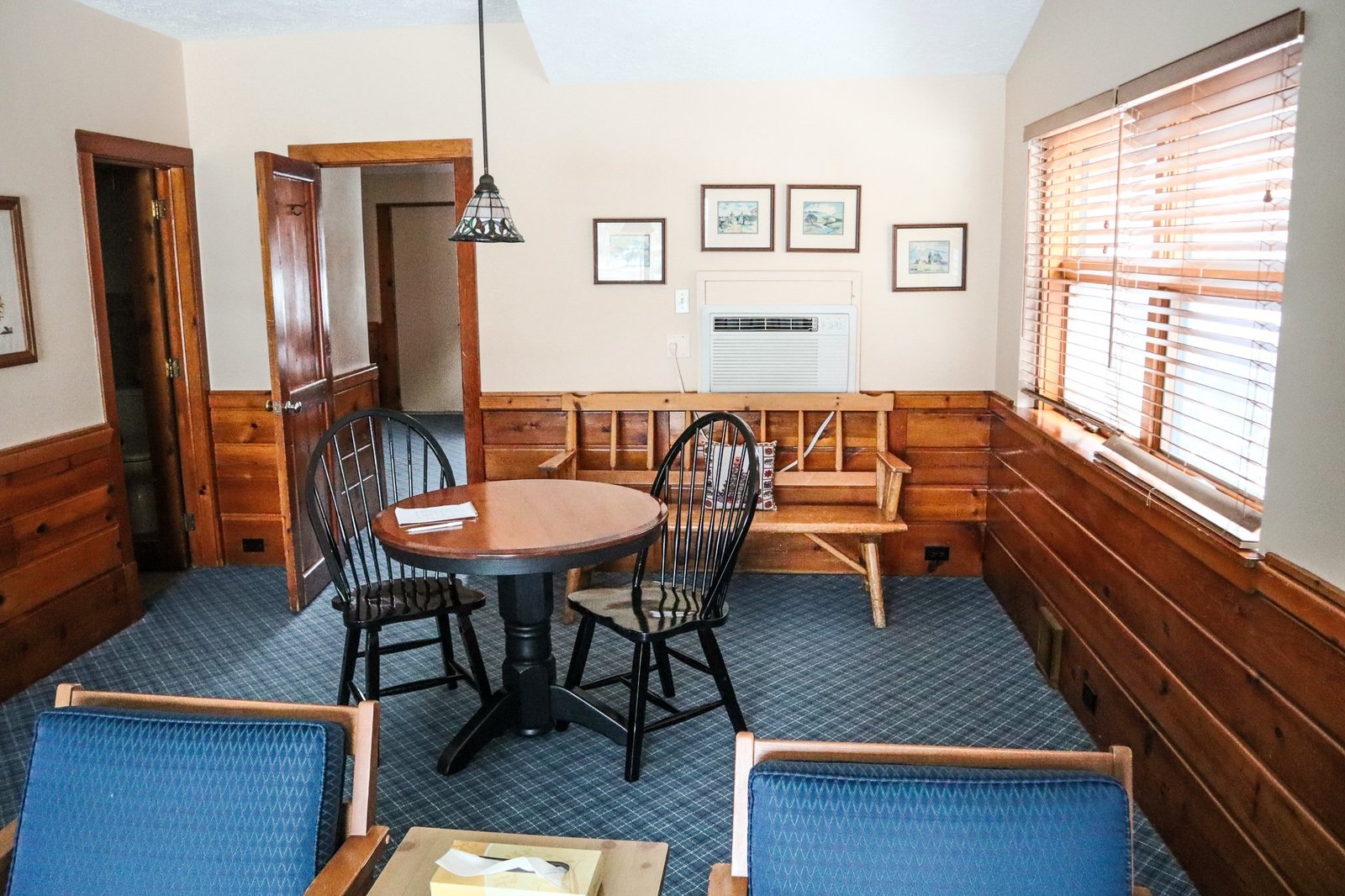 Spacious chalet living area featuring a round table with black chairs, wooden bench, air conditioning unit, and large windows with blinds, highlighting the cozy ambiance of Otsego Resort accommodations.