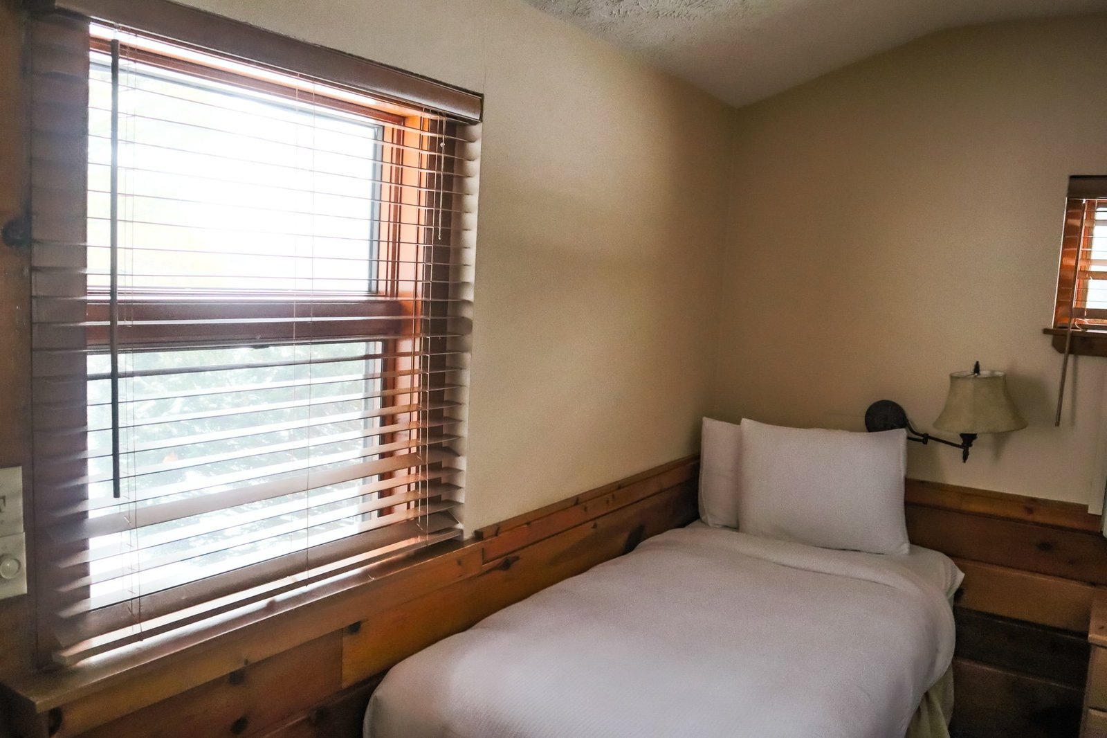 Cozy chalet bedroom featuring a single bed, wooden accents, window with blinds, and ambient lighting, showcasing the relaxing accommodations at Otsego Resort.