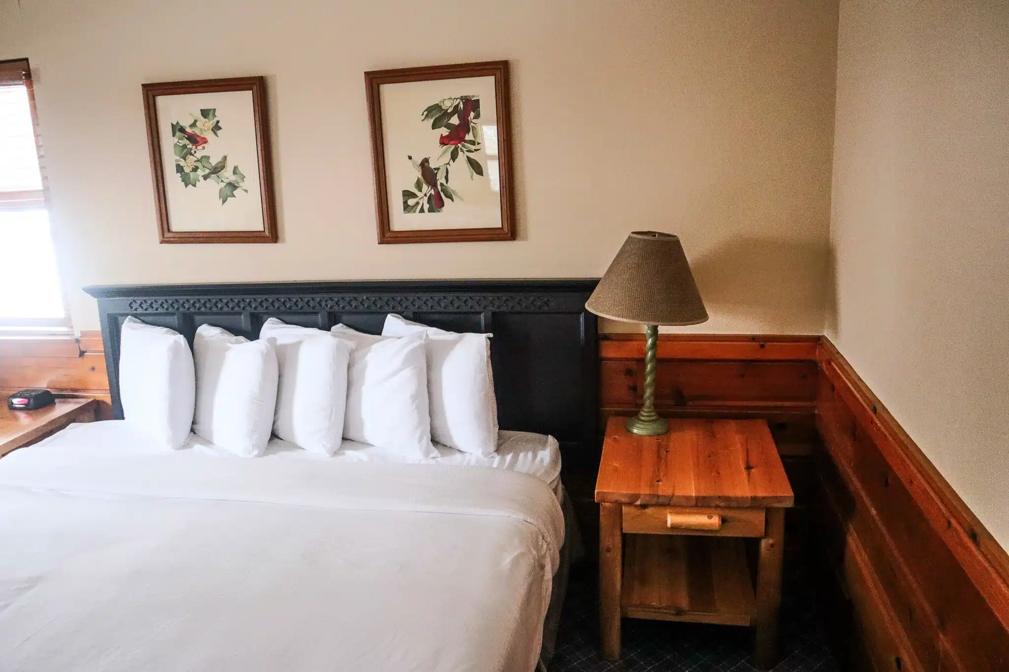 Cozy chalet bedroom featuring a king-sized bed with multiple pillows, wooden nightstand with lamp, and nature-themed artwork on the walls, reflecting the spacious accommodations at Otsego Resort.