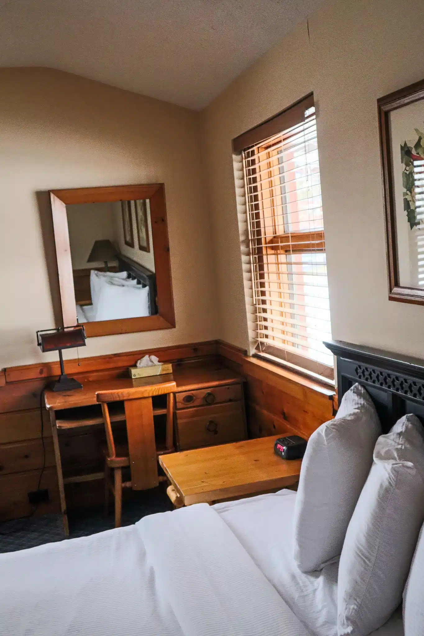 Cozy chalet interior featuring a bed with white linens, wooden furniture including a desk and chair, a mirror, and a window with blinds, showcasing the inviting atmosphere of Otsego Resort accommodations.