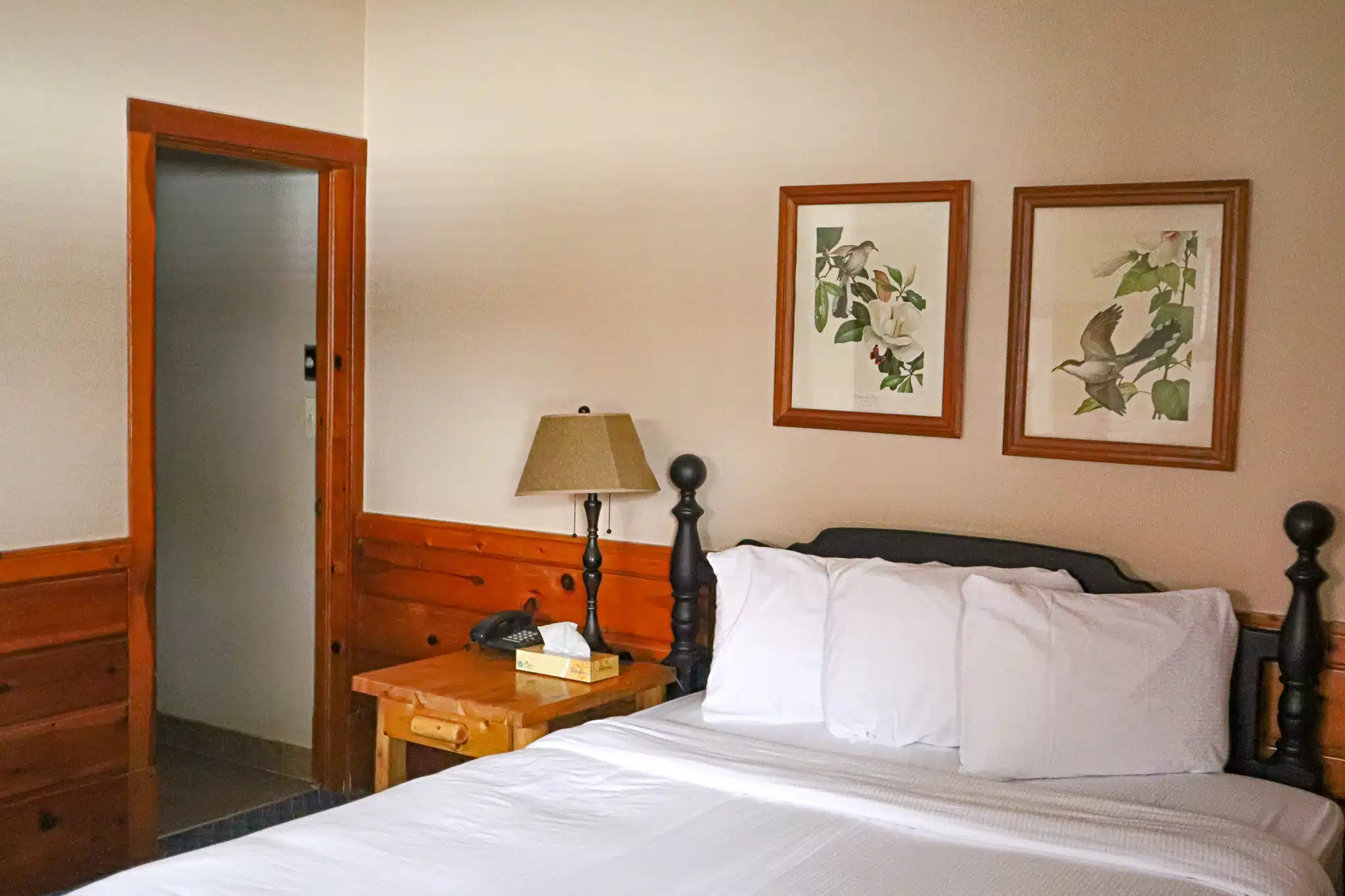 Cozy chalet interior featuring a queen bed with white linens, bedside lamp, wooden furniture, and nature-themed artwork on the walls, ideal for guests at Otsego Resort.
