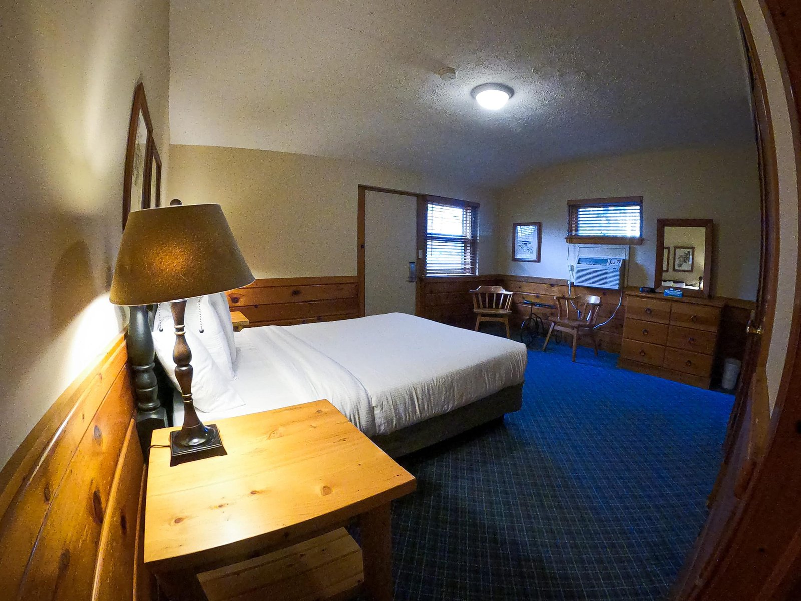 Spacious chalet interior featuring a queen bed, wooden furniture, a lamp, seating area with chairs, and large windows, designed for comfort at Otsego Resort.