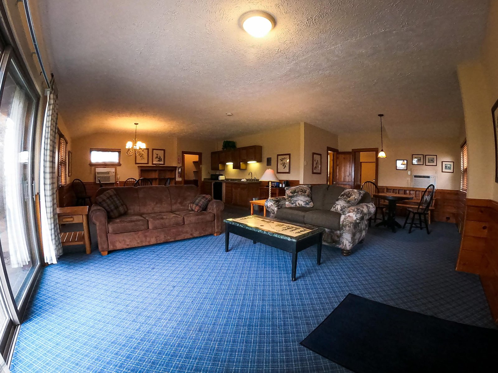 Spacious living room of a two-bedroom chalet at Otsego Resort, featuring a brown sofa, floral couch, kitchenette, dining area, and large windows with natural light.