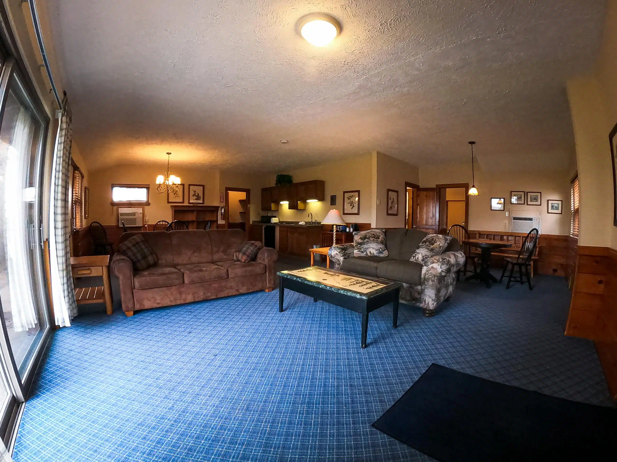 Spacious living room of a two-bedroom chalet at Otsego Resort, featuring a brown sofa, floral couch, kitchenette, dining area, and large windows with natural light.