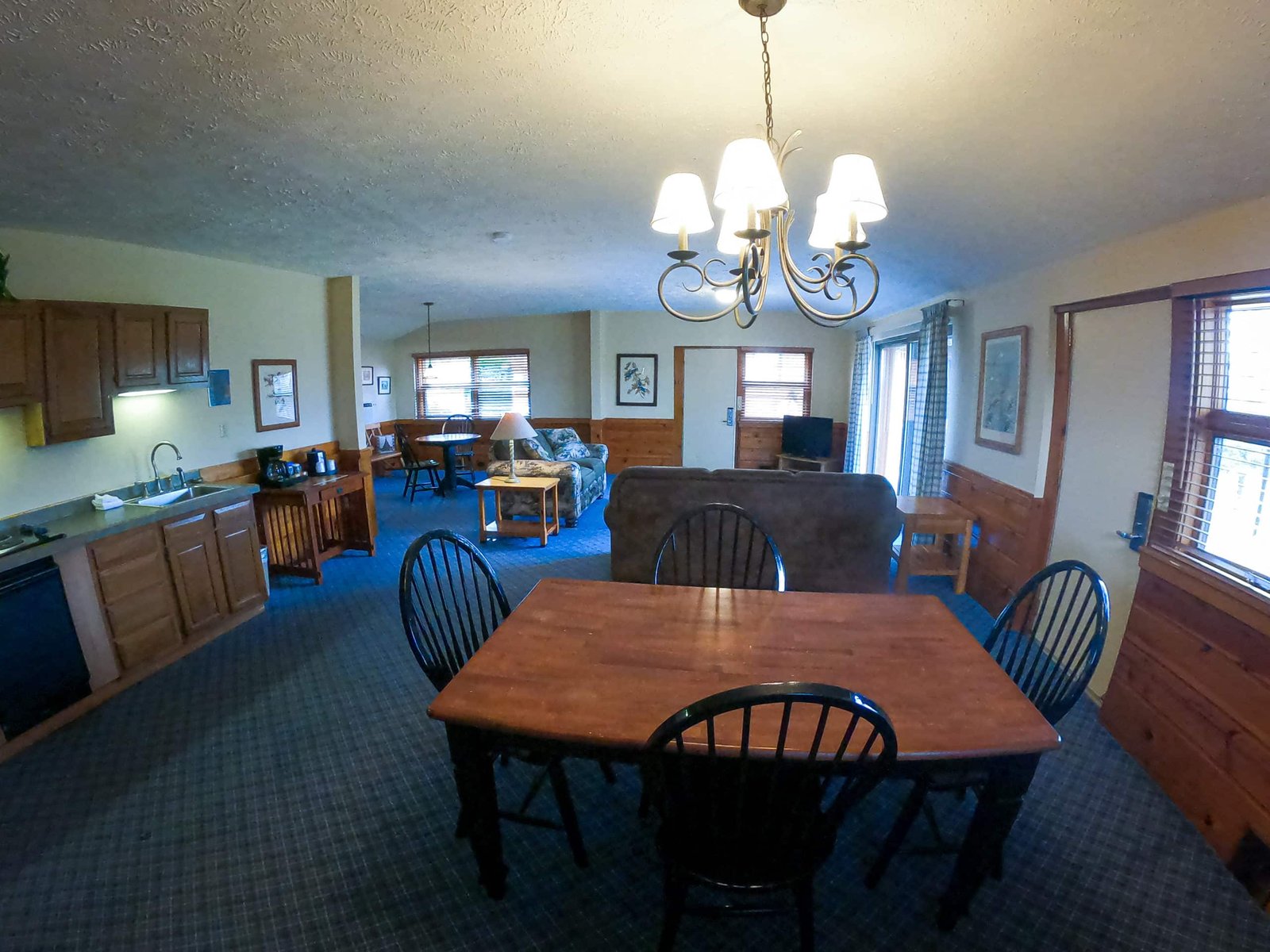 Spacious chalet interior featuring a kitchenette, dining area with wooden table and chairs, living room with sofa, and large windows providing natural light, ideal for family accommodations at Otsego Resort.