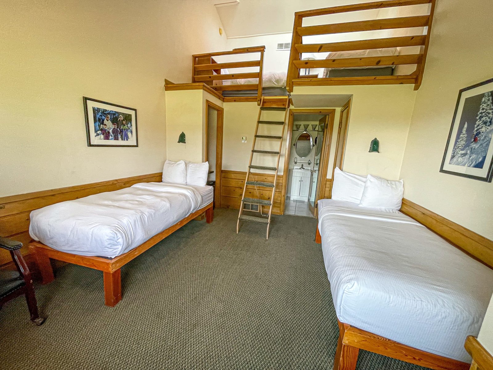 Loft Lodge room at Otsego Resort featuring two twin beds, a wooden ladder leading to a loft area, and a full bath visible in the background, showcasing cozy northern Michigan accommodations.