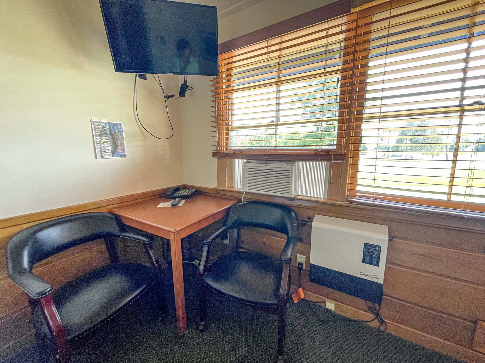 Cozy workspace in Loft Lodge room featuring a wooden table, two black chairs, a wall-mounted TV, air conditioning unit, and natural light from window blinds, reflecting the comfortable amenities offered at Otsego Resort.