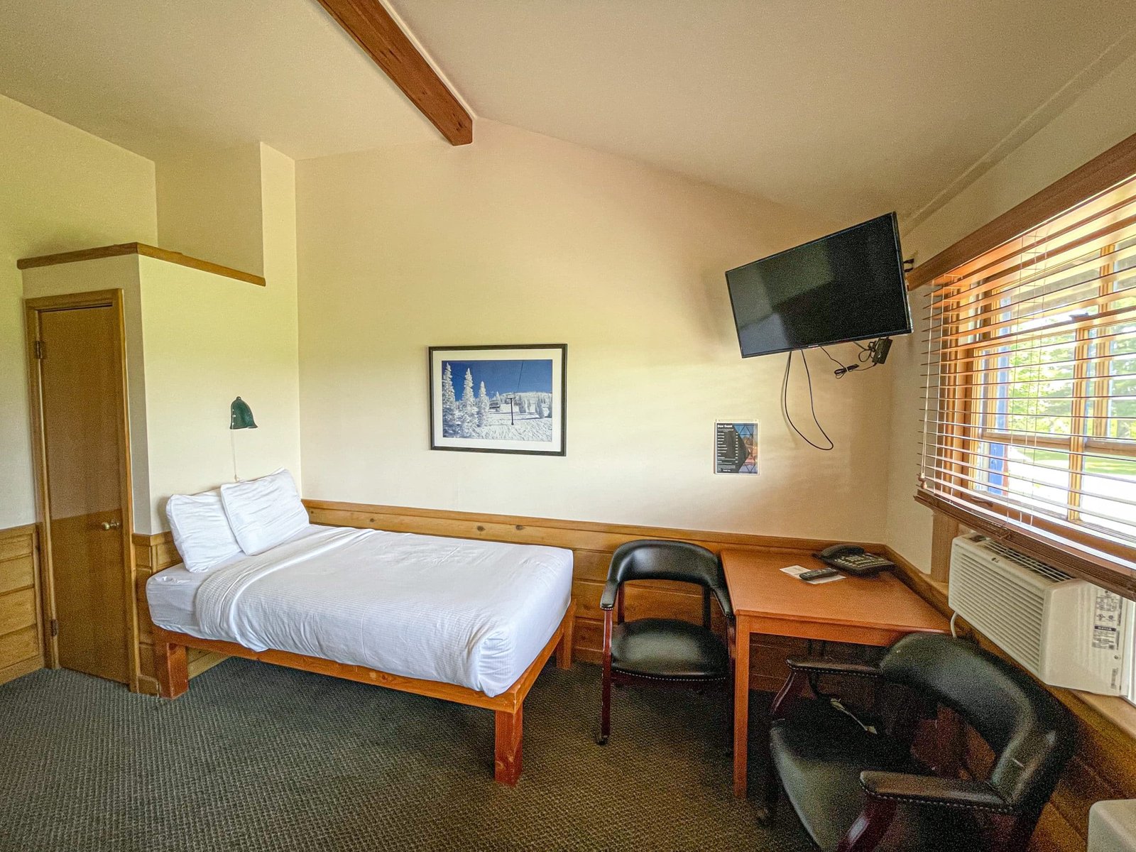 Cozy hotel room at the Loft Lodge featuring a twin bed, workspace with table and chairs, wall-mounted television, and natural light from window with blinds, reflecting the inviting atmosphere of northern Michigan accommodations.