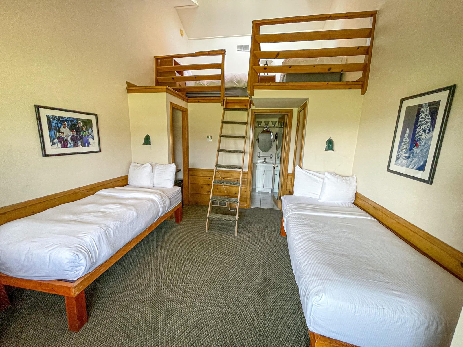Loft Lodge hotel room featuring two twin beds, a lofted sleeping area, wooden furnishings, and a full bath visible through an open door, highlighting comfort and convenience for guests at Otsego Resort.