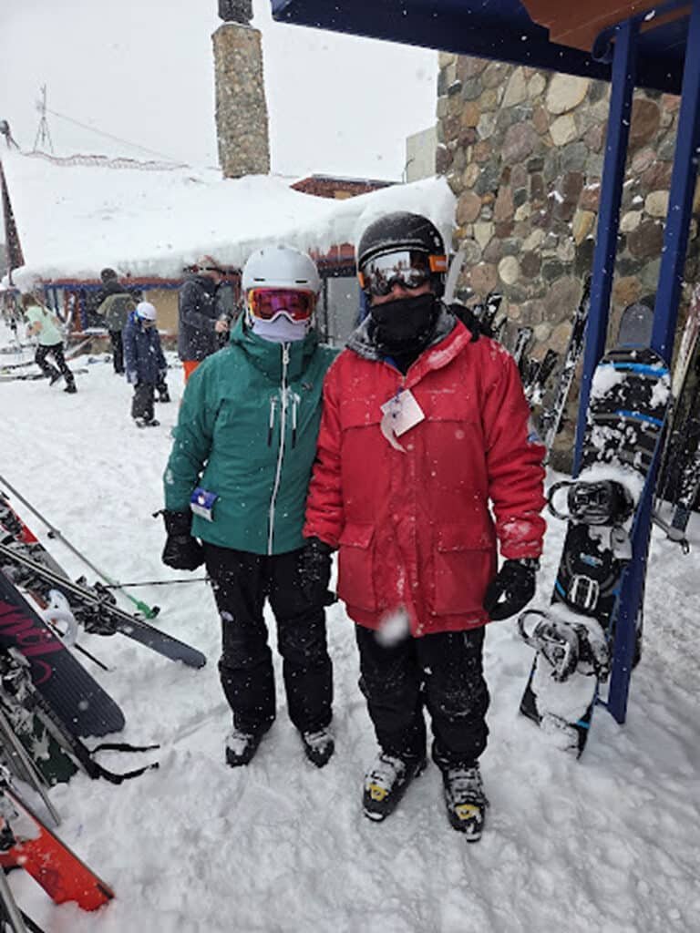 Two skiers in winter gear smiling in snowy outdoor setting, with skis and snowboards nearby, representing personalized ski lessons at Otsego Resort.