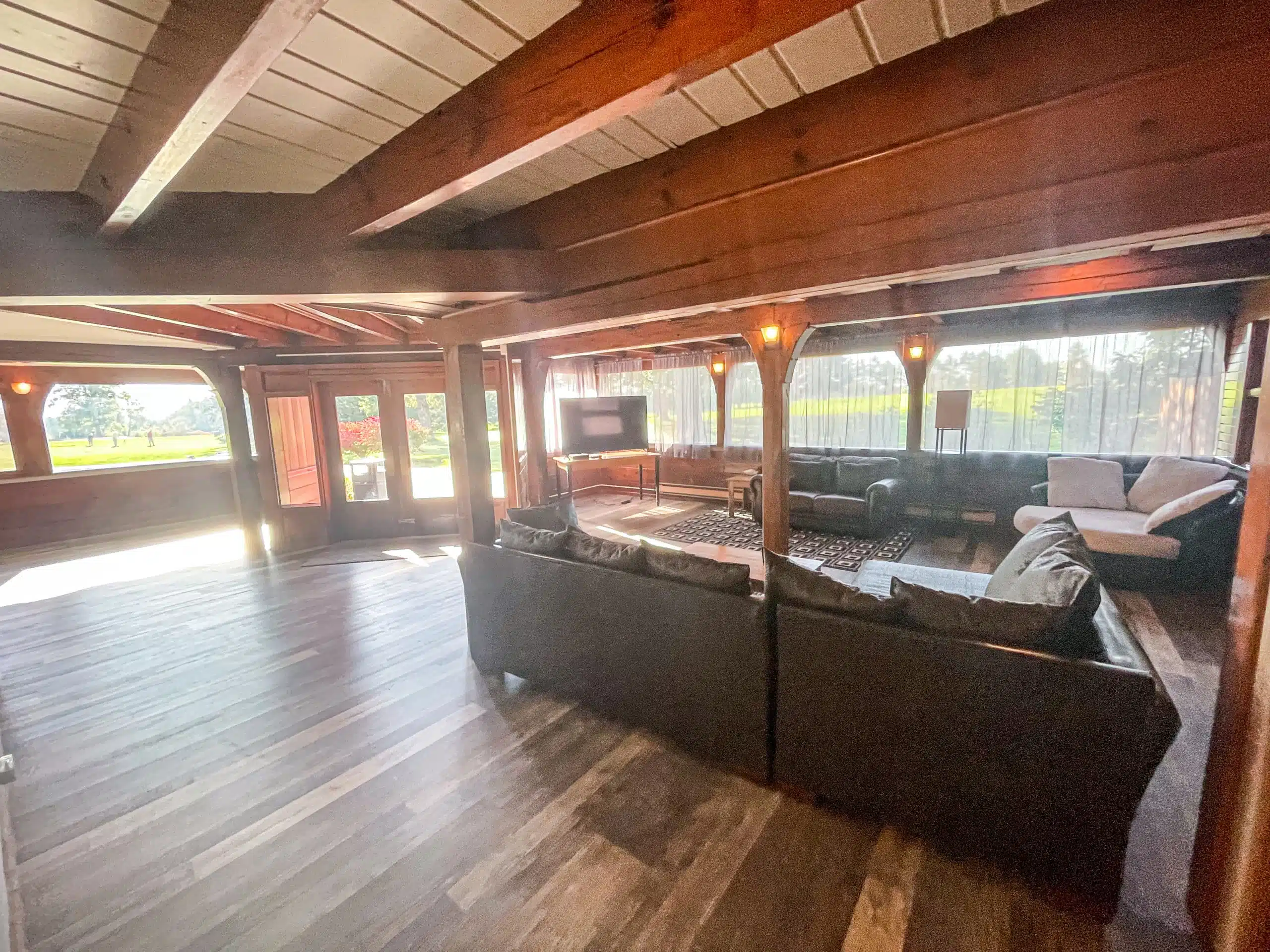 Large living room in the Classic Lodge at Otsego Resort, featuring dark leather couches, a flat screen TV, wooden beams, and expansive windows overlooking the golf course.