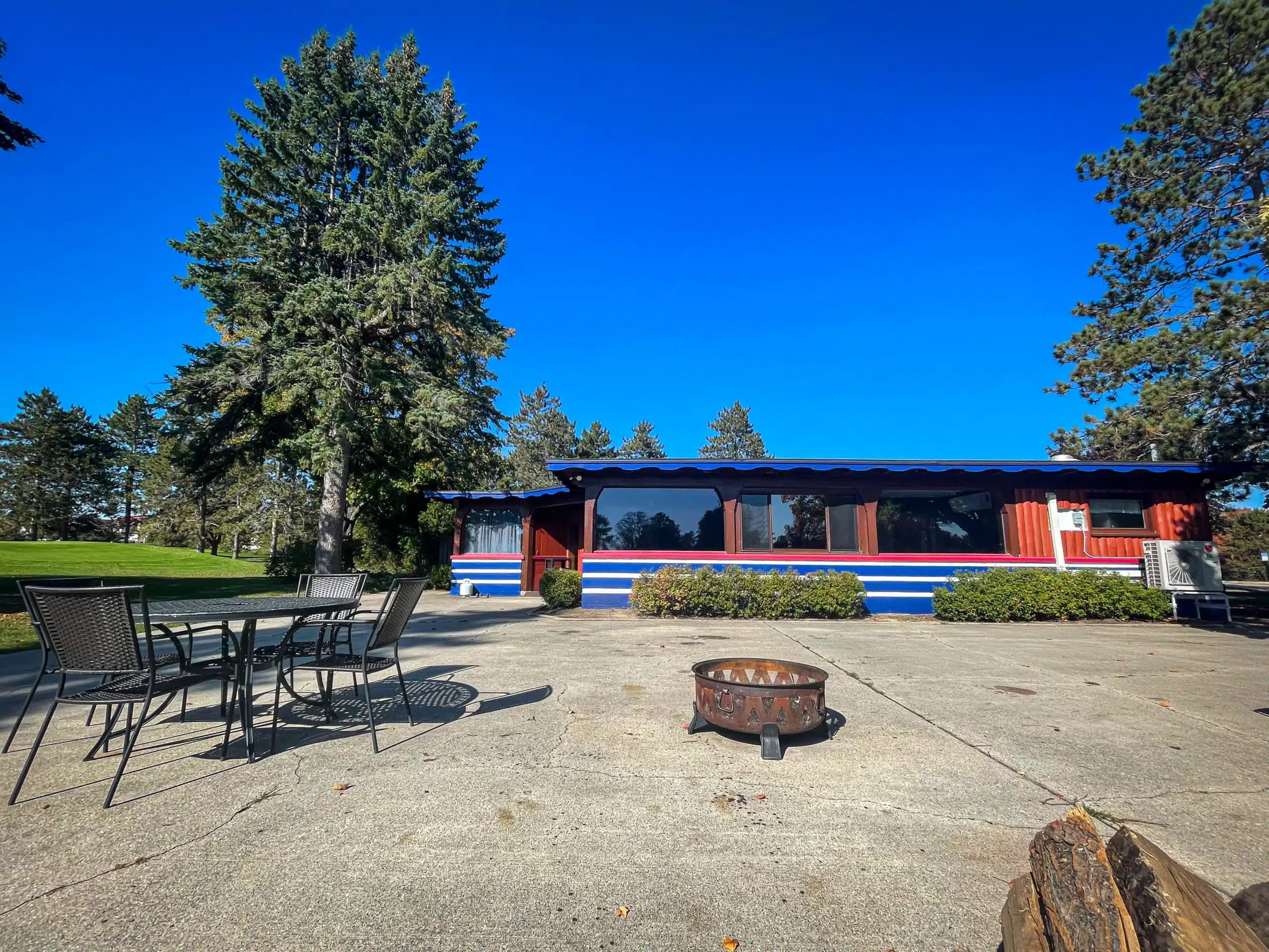 Exterior view of The Classic Lodge at Otsego Resort, featuring a renovated building with blue and red accents, surrounded by trees, a patio with a table and chairs, and a fire pit, located near the 18th hole of the golf course.