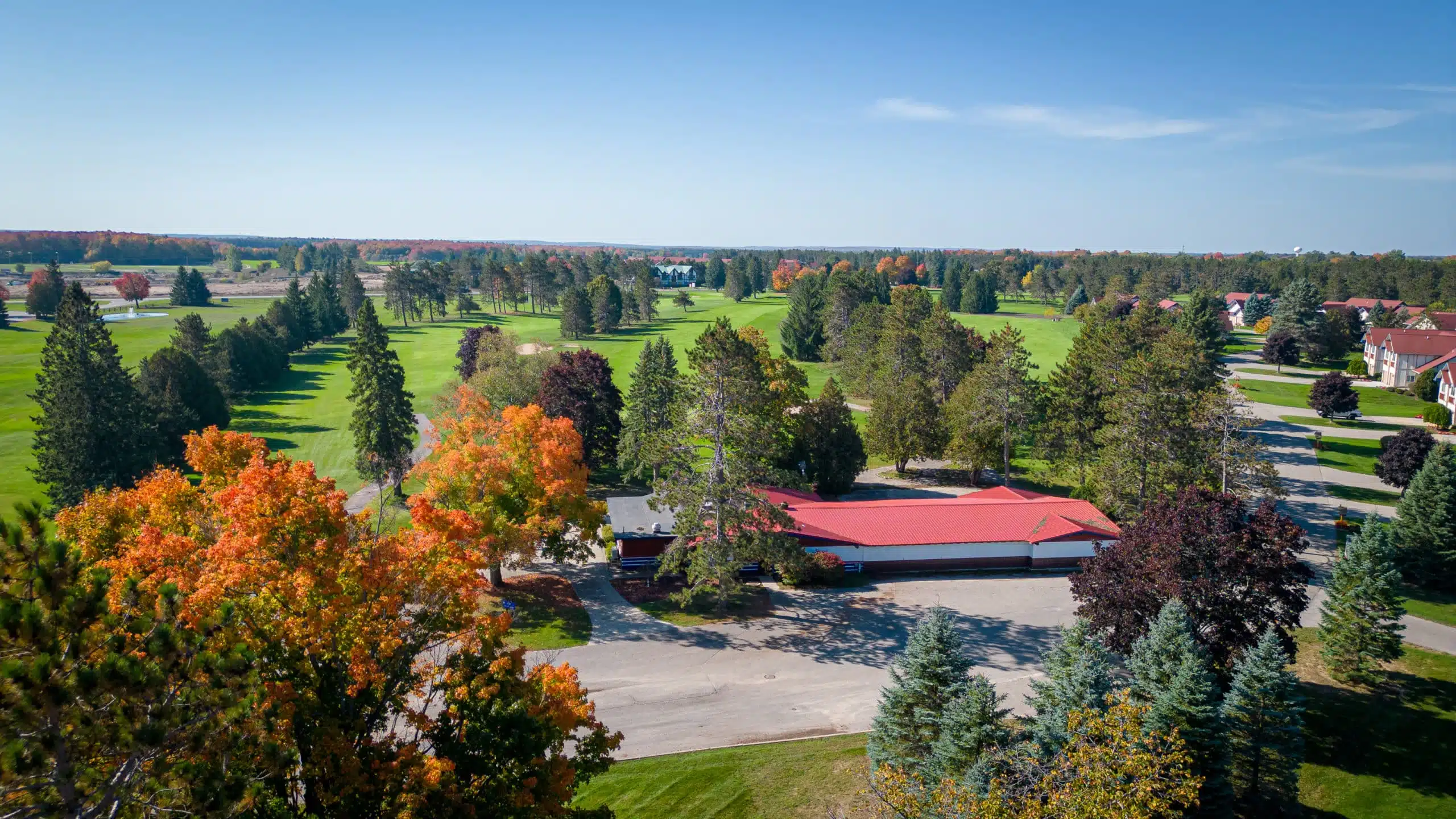 Aerial view of The Classic Lodge at Otsego Resort, surrounded by vibrant autumn foliage and the 18th hole of the golf course, showcasing the lodge's red roof and nearby residential accommodations.
