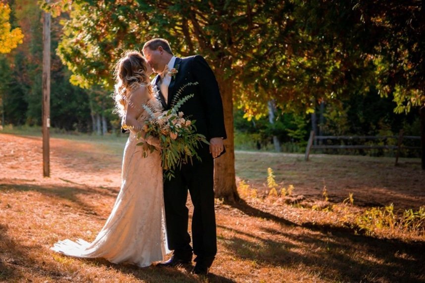 Bride and groom sharing a kiss in a scenic outdoor setting, surrounded by autumn foliage, showcasing a romantic moment ideal for weddings at Otsego Resort.