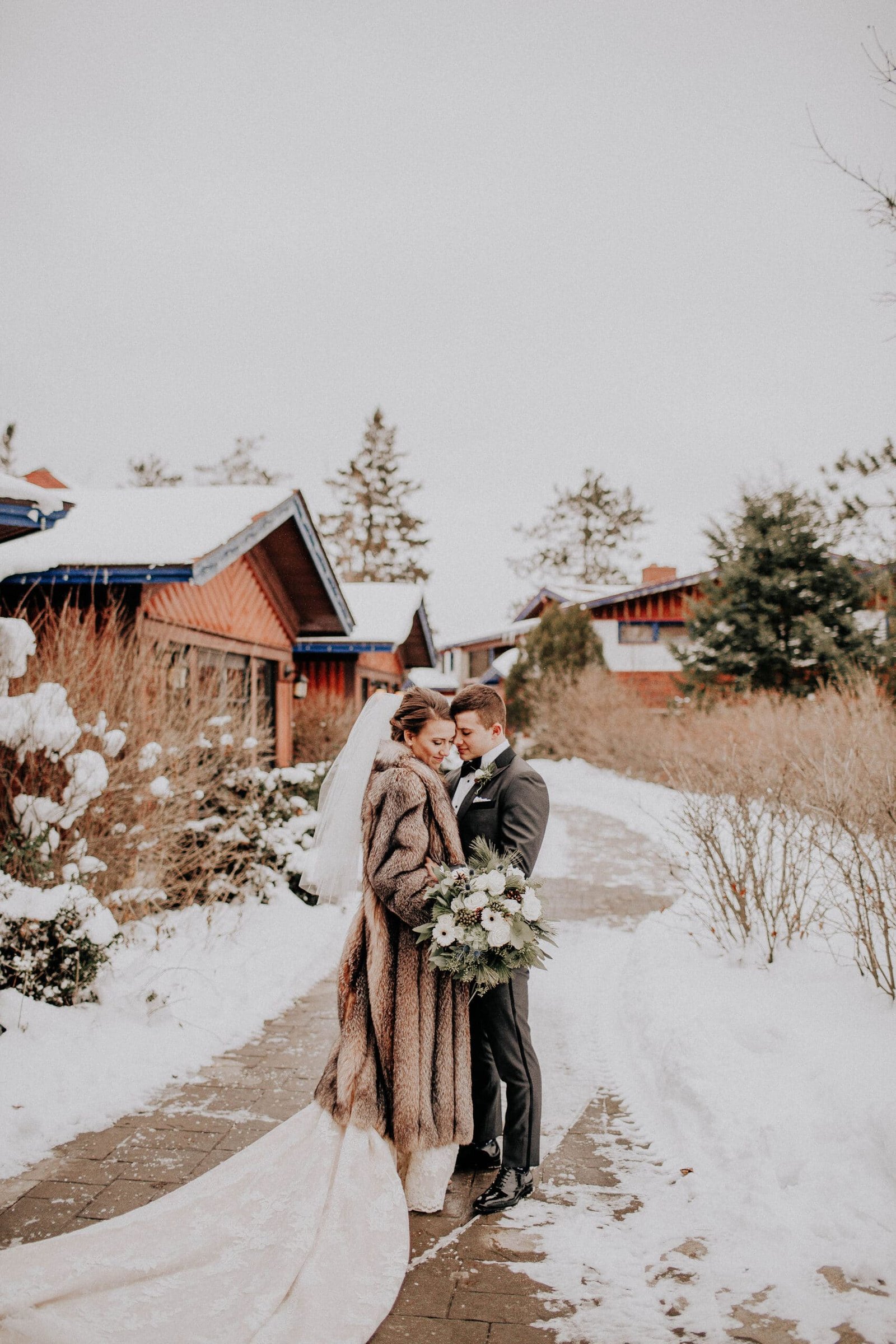 Bride and groom embracing in winter setting, wearing elegant wedding attire, surrounded by snow-covered cabins and trees at Otsego Resort.