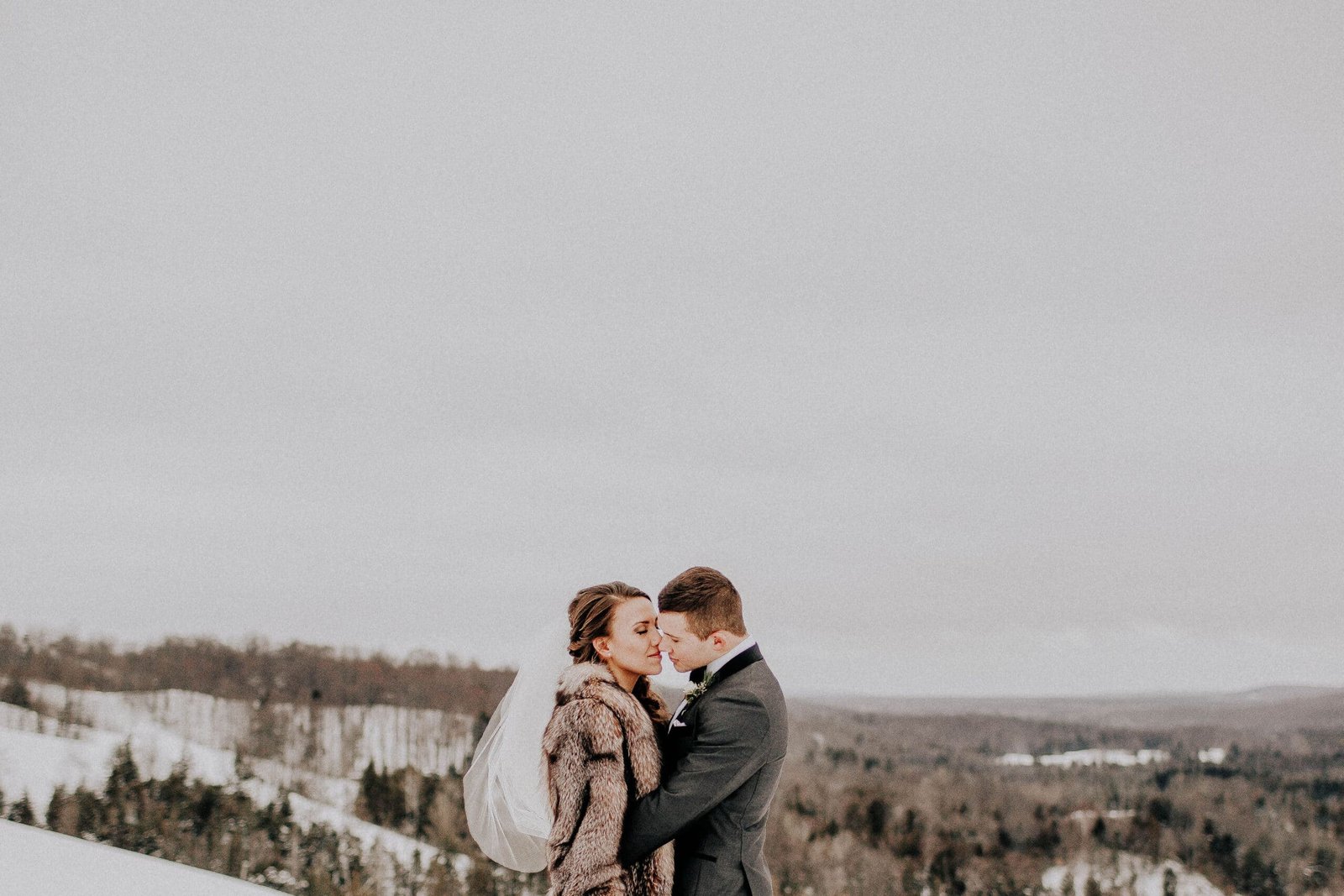 Couple embracing in winter attire with snowy landscape of Sturgeon River Valley in the background, capturing a romantic moment at Otsego Resort.