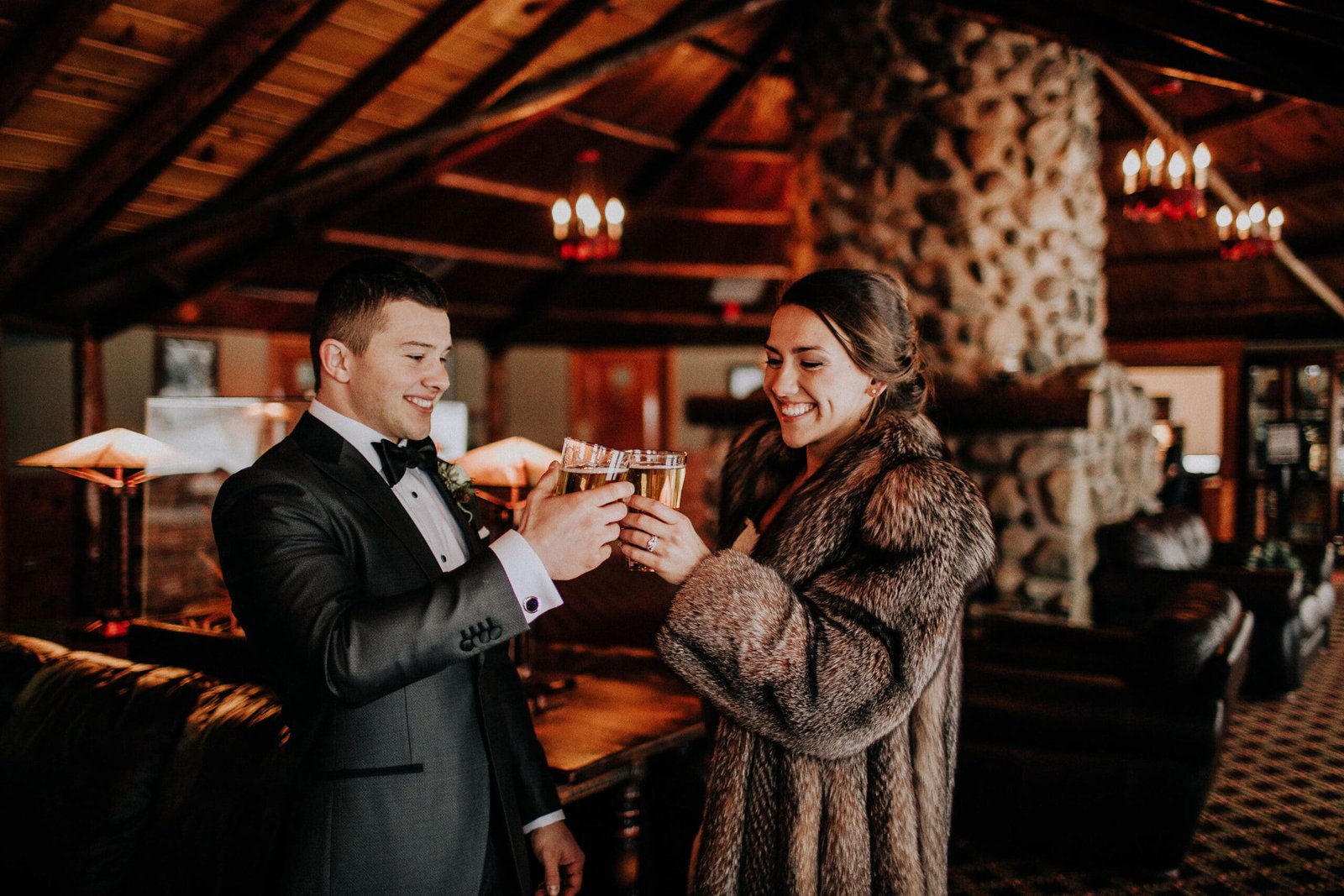 Couple toasting with drinks in a cozy lodge setting at Otsego Resort, celebrating a special occasion amid rustic decor and warm lighting.