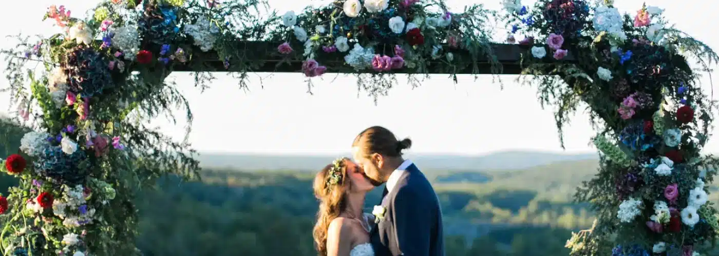 Couple kissing under a floral wedding arch with scenic valley views, capturing a romantic outdoor ceremony at Otsego Resort.