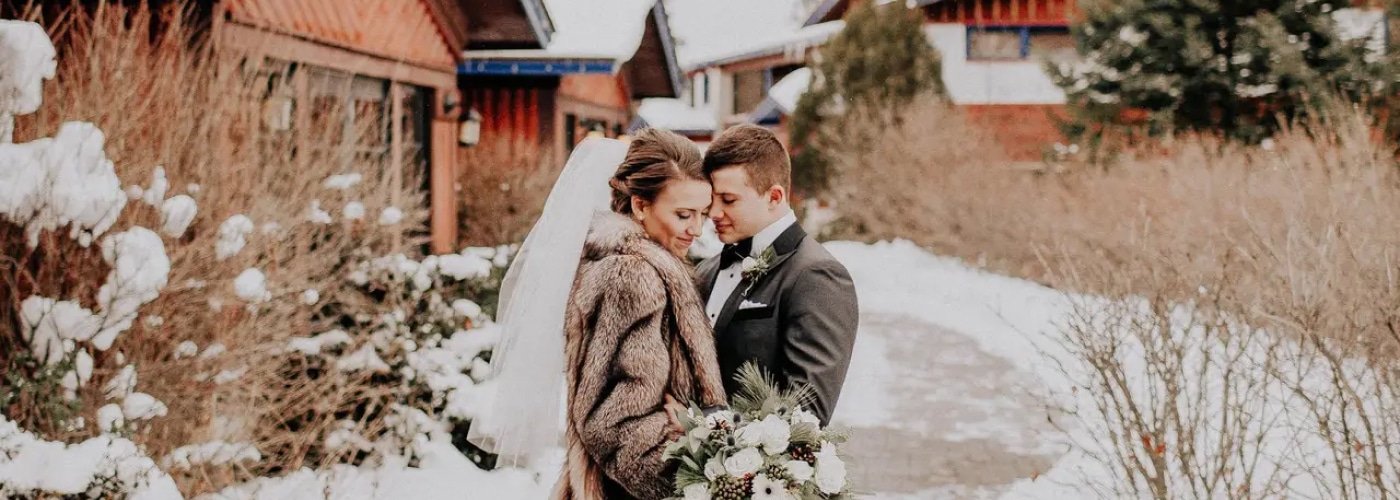 Bride and groom embracing in winter attire, surrounded by snow and rustic lodge backdrop, capturing a romantic moment at Otsego Resort wedding.