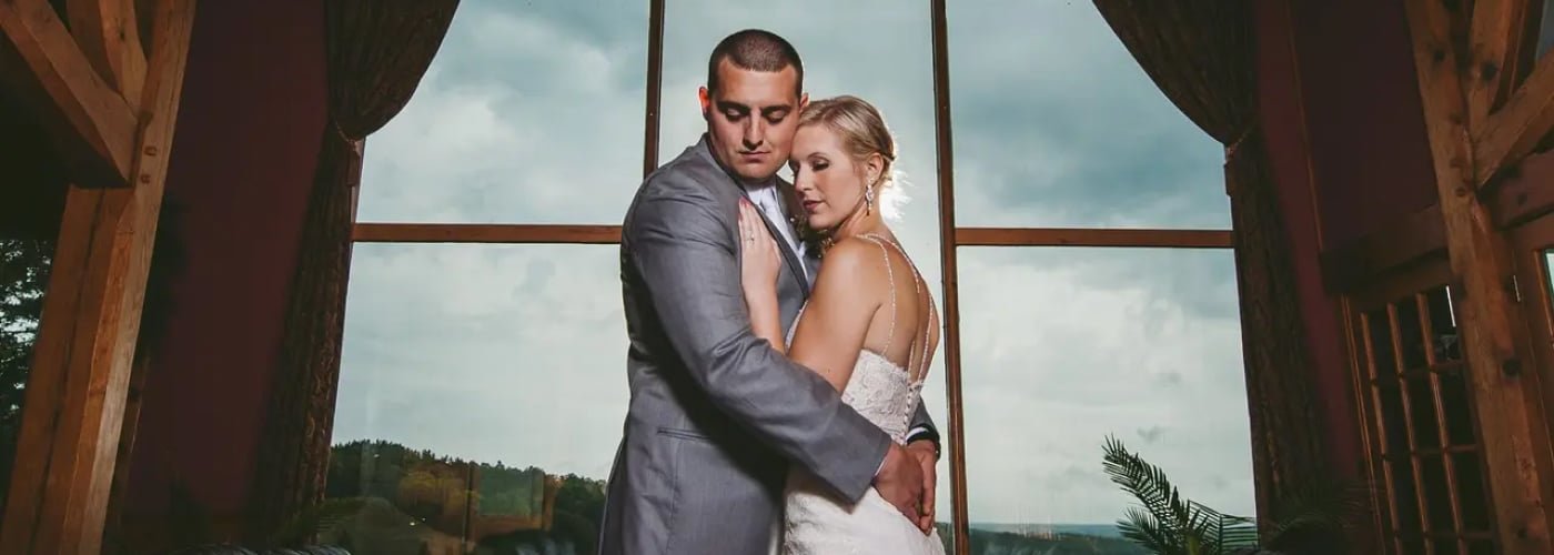 Bride and groom embracing in a romantic pose, framed by large windows showcasing a scenic valley view, within the Bavarian-themed Otsego Resort setting.