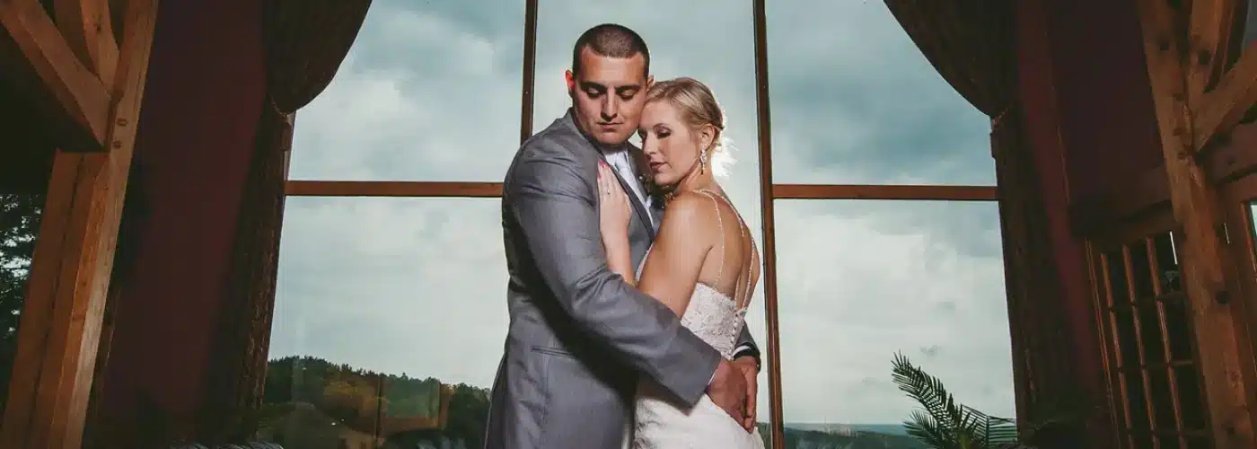 Bride and groom embracing in a romantic pose, framed by large windows showcasing a scenic valley view, within the Bavarian-themed Otsego Resort setting.