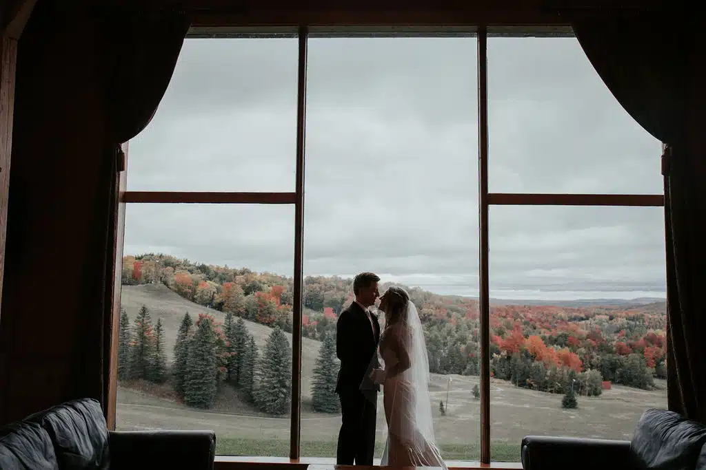 Couple sharing a kiss in front of large window with fall valley views, showcasing Otsego Resort's romantic wedding setting.